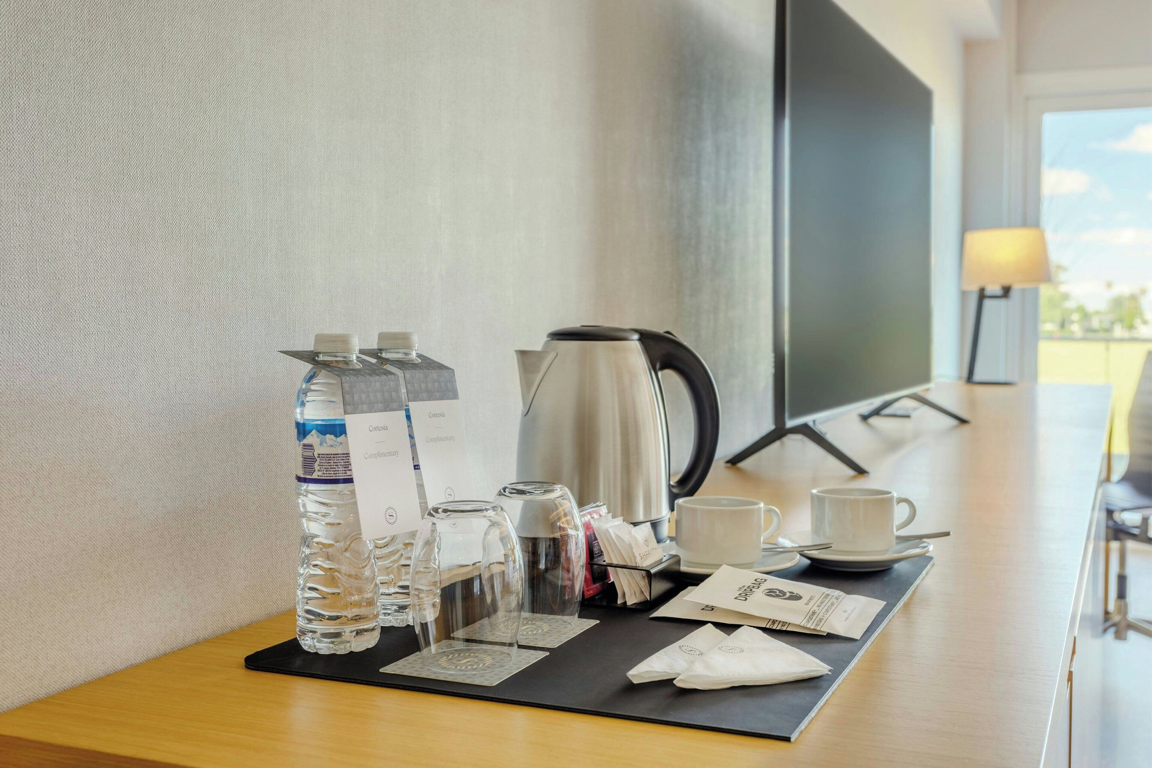 A hotel room with a wooden desk, a kettle, a water bottle, a glass, a cup, and a tray with napkins and a menu.