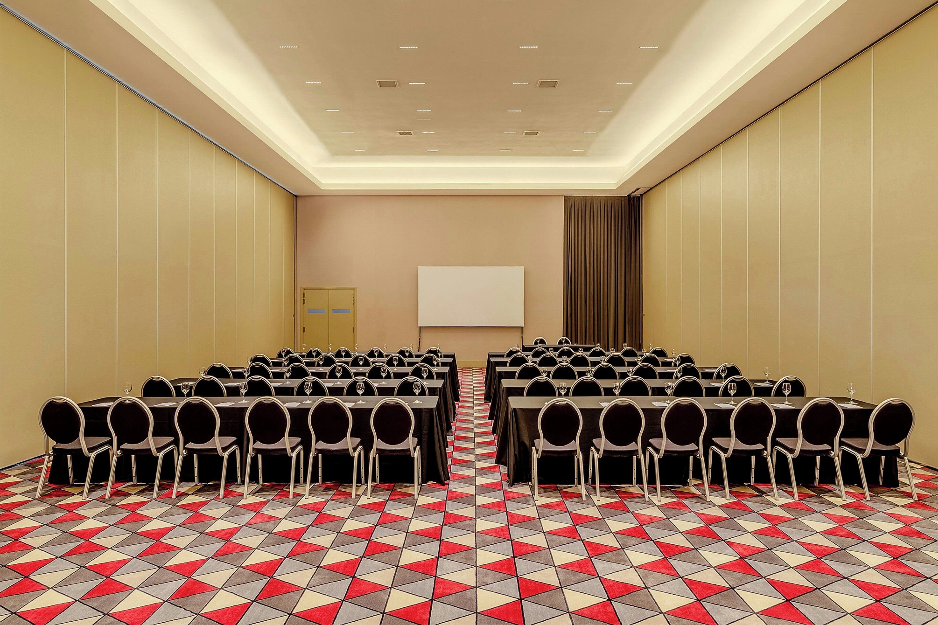 Un salón de conferencias con filas de mesas y sillas, una pantalla de proyector y una alfombra estampada.