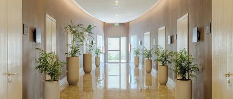 Un pasillo de hotel con suelo de mármol pulido, plantas en macetas grandes y puertas a ambos lados.