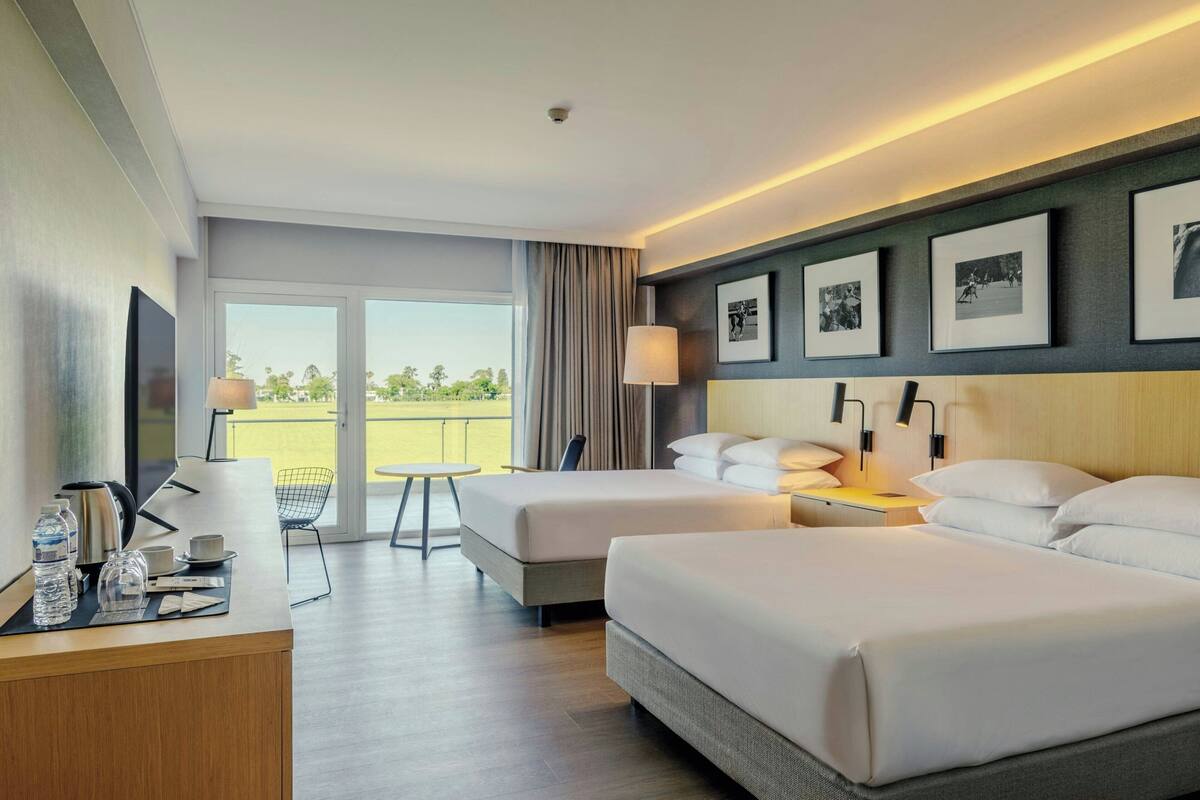 A hotel room with two beds, a desk with a chair, a TV, and a balcony with a view.