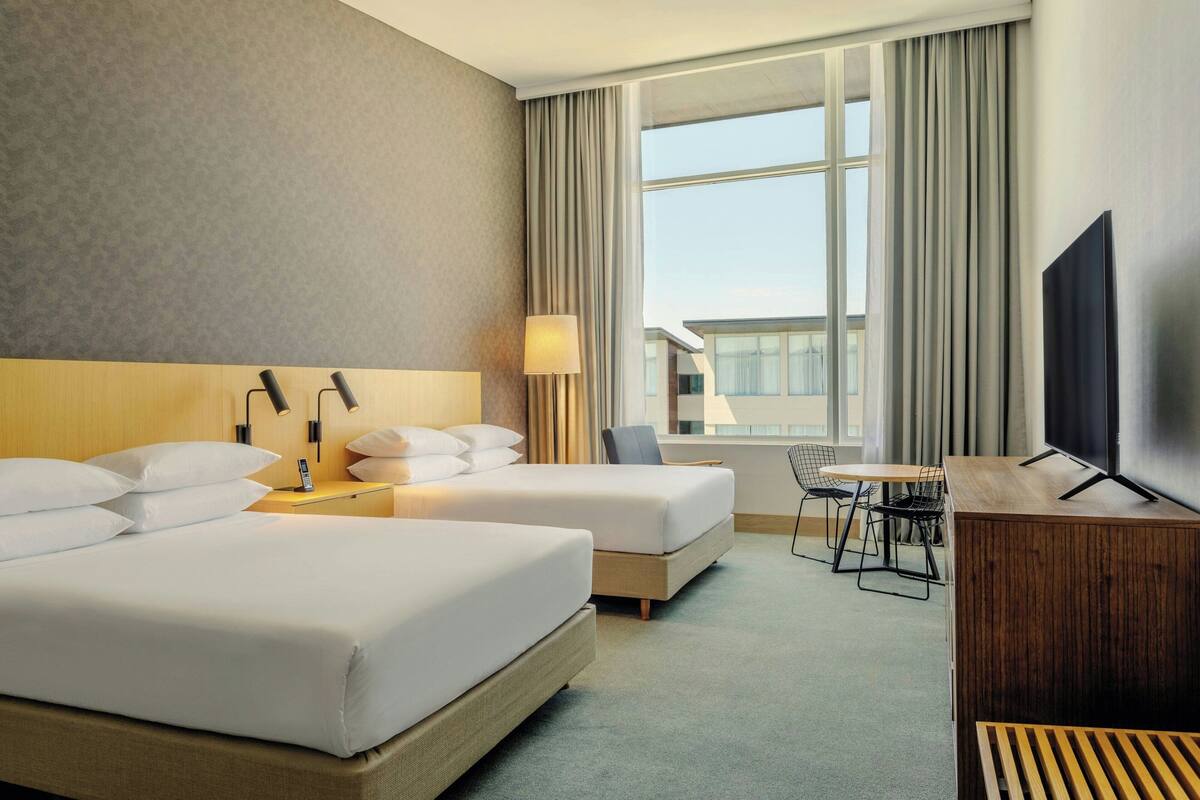 A hotel room with two beds, a desk with a lamp, a TV, and a view of the city.