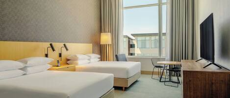 A hotel room with two beds, a desk with a lamp, a TV, and a view of the city.