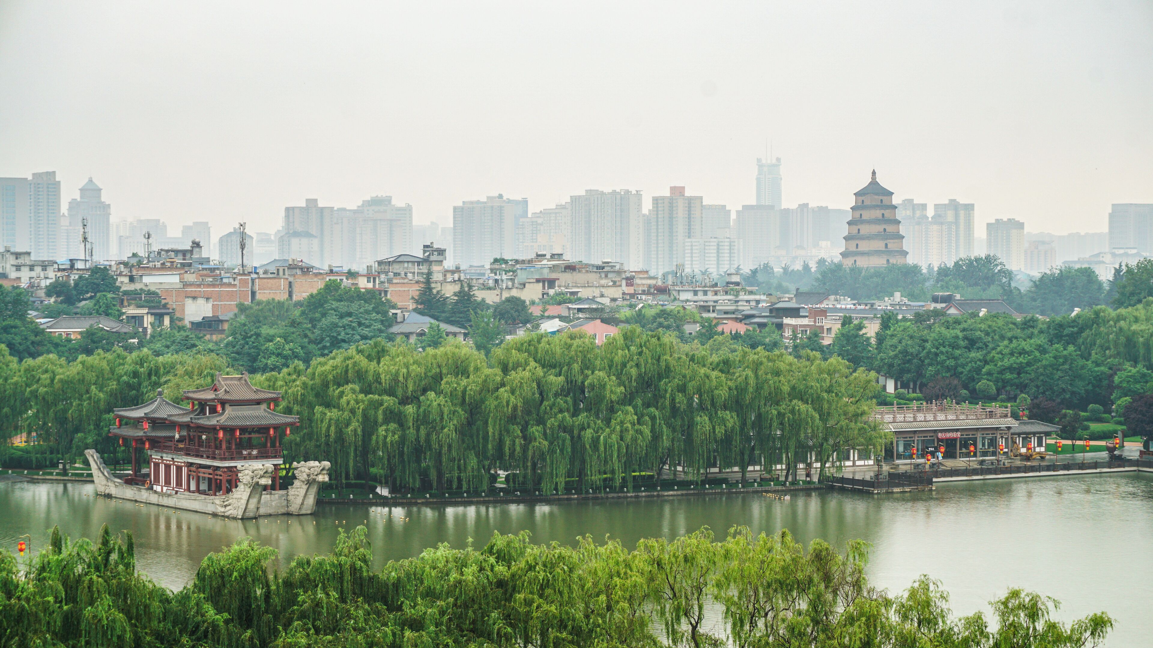 Een traditioneel Chinees paviljoen aan een meer, met een stadsgezicht op de achtergrond.