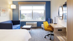 A hotel room with a bed, a blue sofa, a yellow chair, a desk, and a large window with a view of trees and buildings. - Holiday Inn Express & Suites Austin South by IHG (Austin)