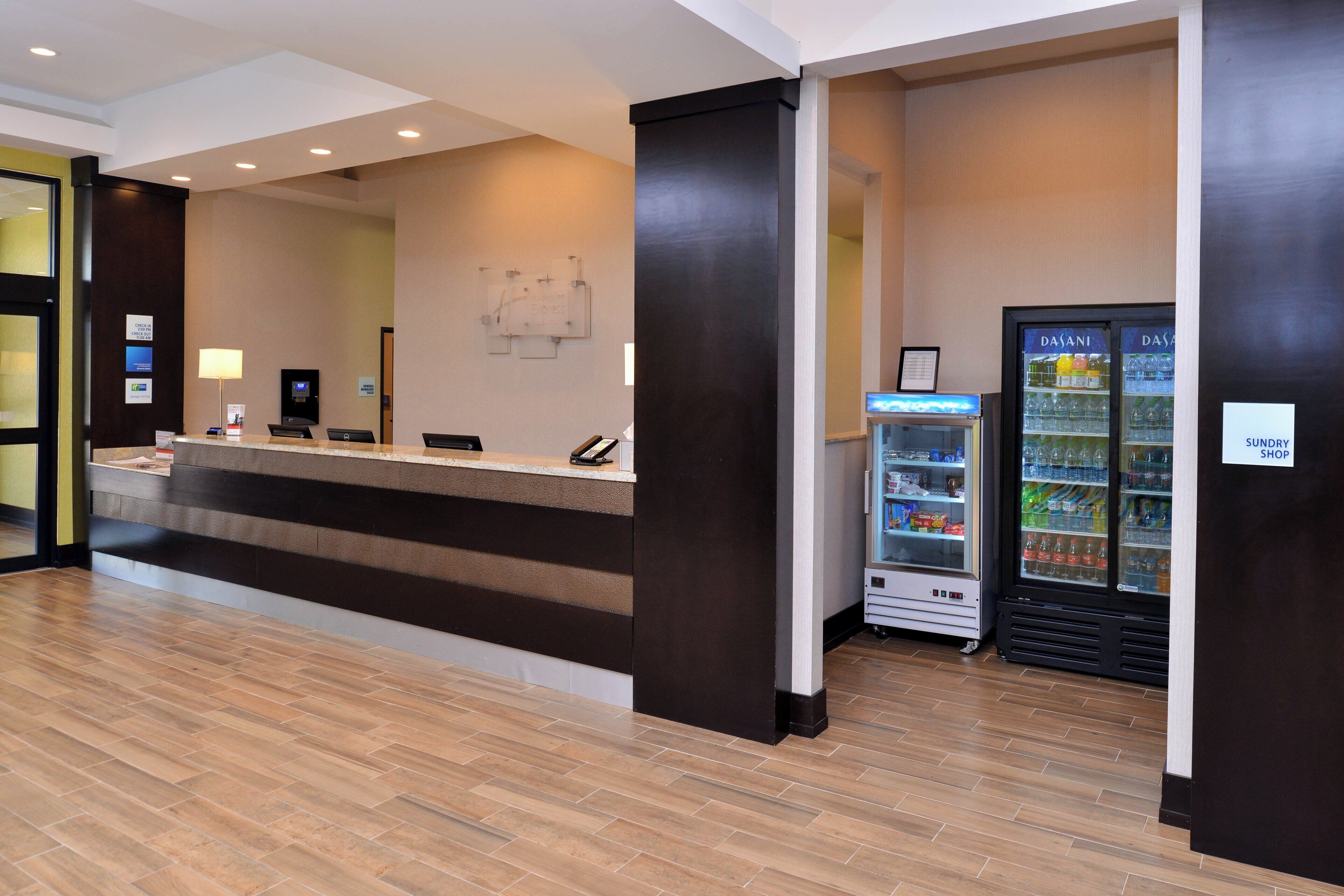 A hotel reception area with a reception desk, a digital check-in kiosk, a refrigerator vending machine, and a sign indicating a sundry shop.