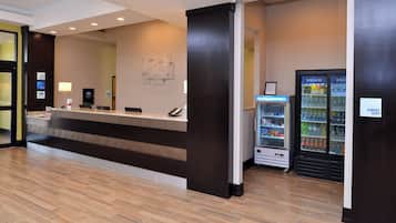 A hotel reception area with a reception desk, a digital check-in kiosk, a refrigerator vending machine, and a sign indicating a sundry shop.