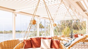 Amplia habitación iluminada por el sol con ventanas amplias que dan a una masa de agua, con mobiliario de mimbre y una silla colgante.