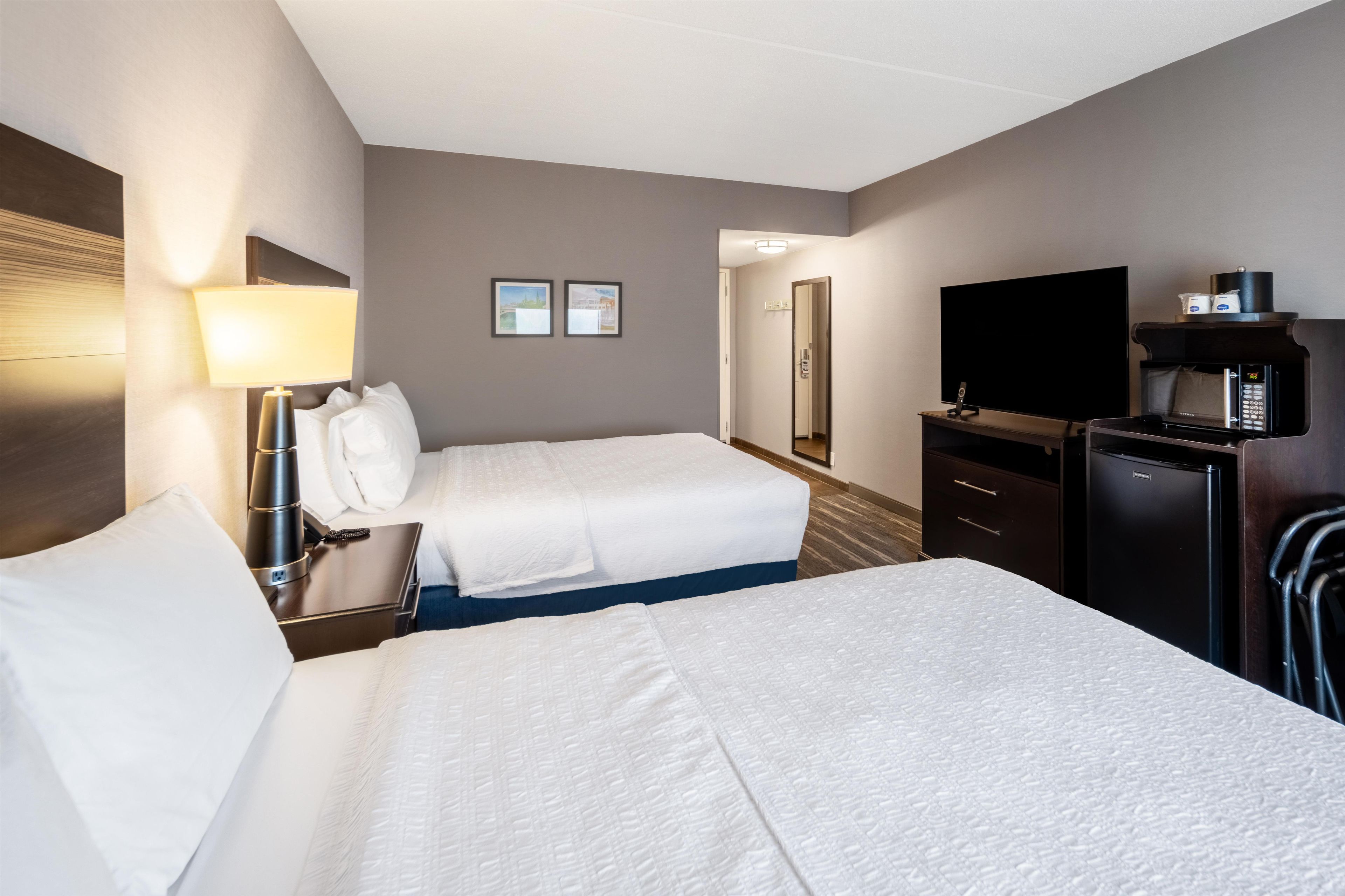 A hotel room with two beds, a desk with a lamp, a television, a minibar, and a mirror.