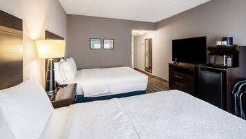 A hotel room with two beds, a desk with a lamp, a television, a minibar, and a mirror.