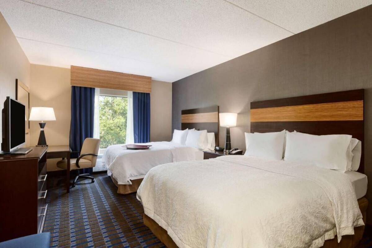 A hotel room with two beds, a desk, a TV, and a window with curtains.