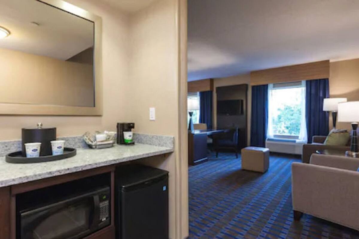 A hotel room with a kitchenette, a large mirror, a flat-screen TV, a seating area with a sofa and armchairs, and a window with curtains.