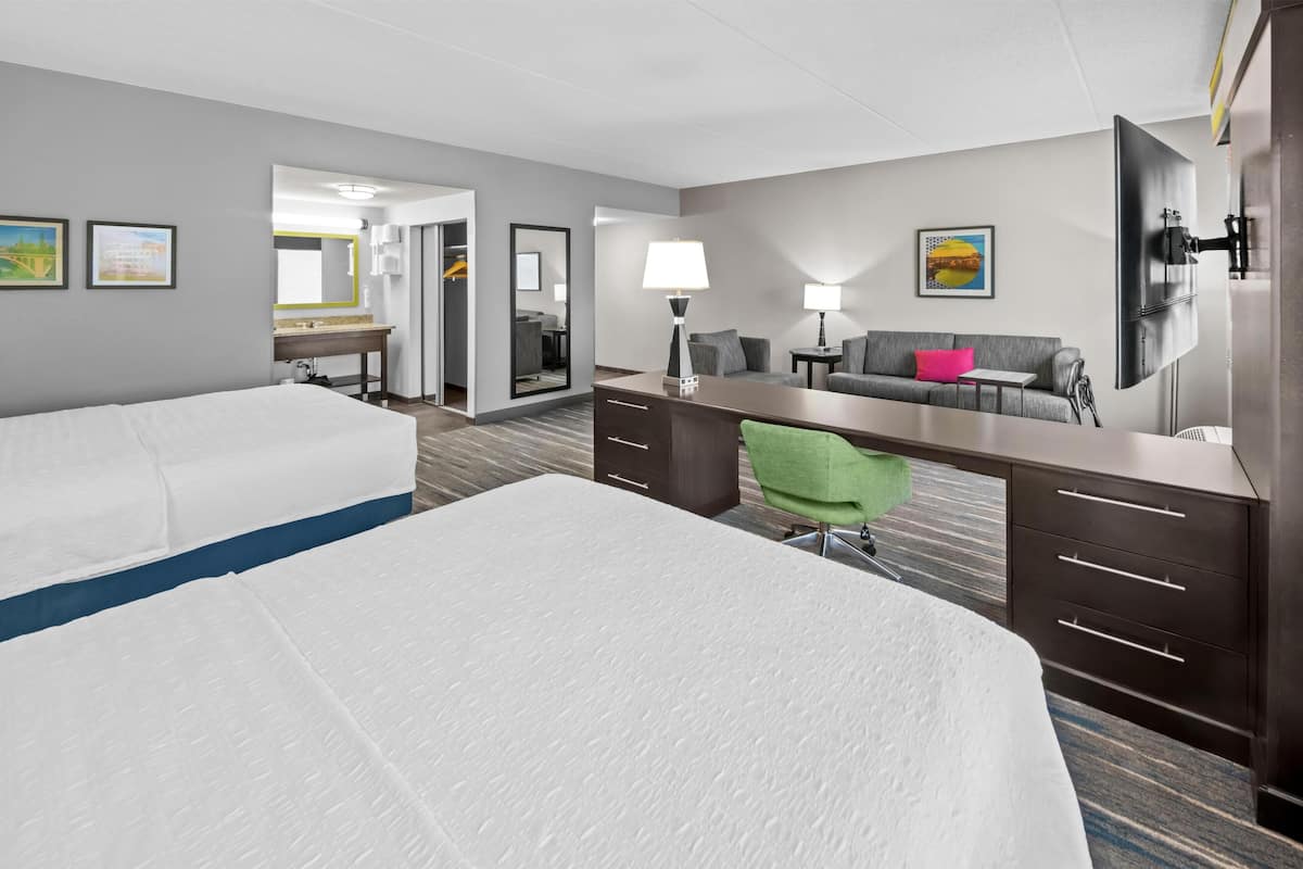 A hotel room with two beds, a desk with a chair, a sofa, a TV, and a mirror.