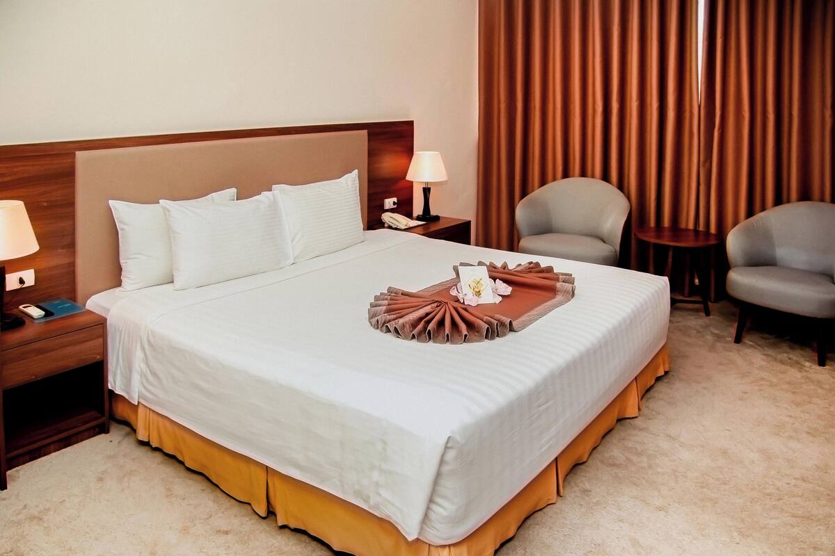 Uma cama bem arrumada com edredom branco e castanho, uma luminária de cabeceira, um telefone e uma pequena mesa com abajur.