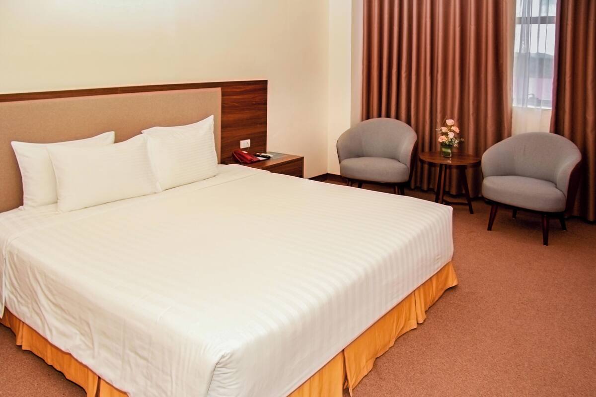 Quarto de hotel com uma cama grande, duas poltronas, uma mesinha de centro com um vaso e uma janela com cortinas.