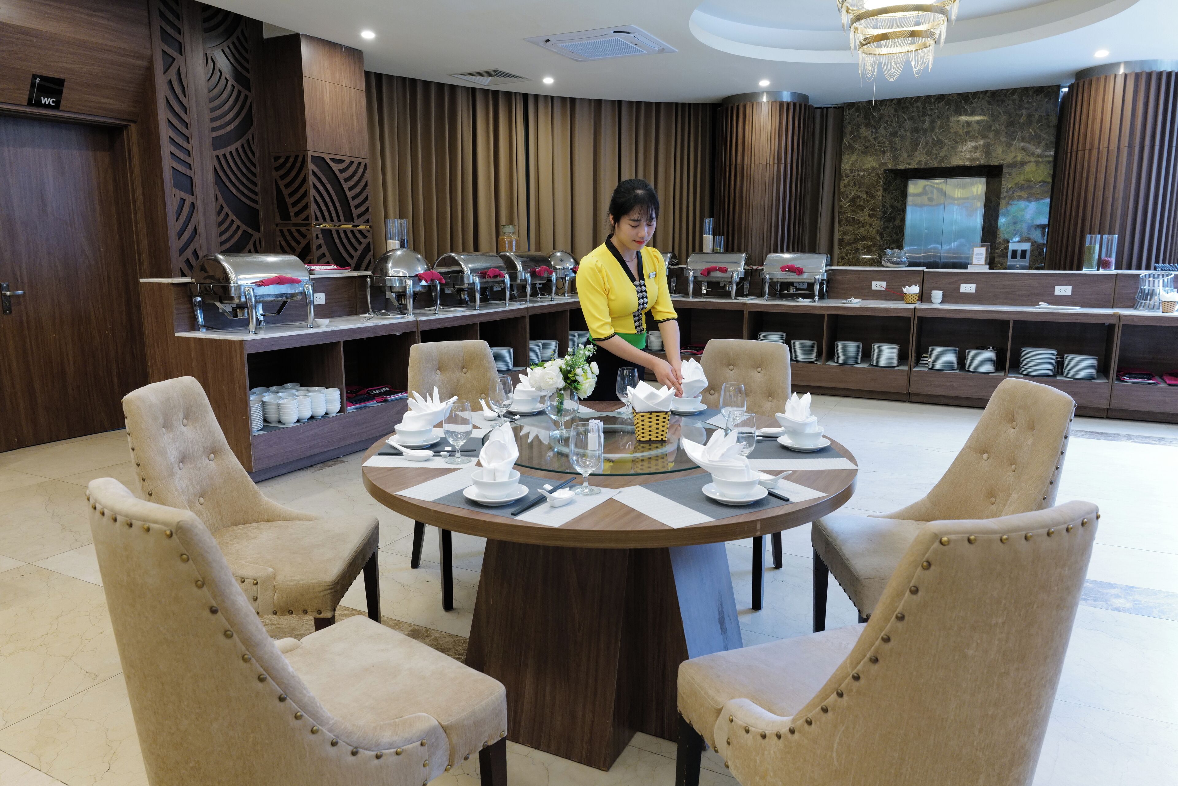 A restaurant setting with a buffet line, neatly arranged tables with white tablecloths, and a staff member in uniform.