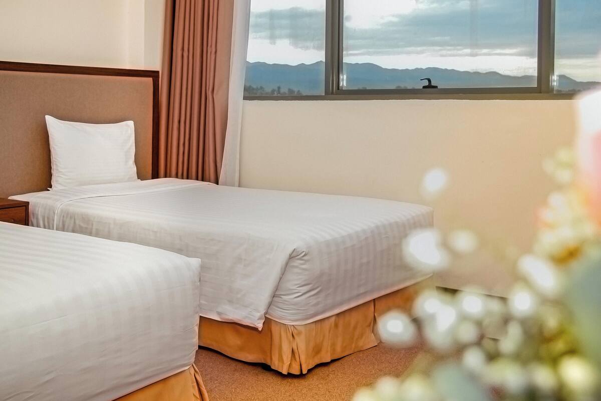 Quarto de hotel com duas camas, uma janela grande com vista para as montanhas e uma mesa de cabeceira com abajur.