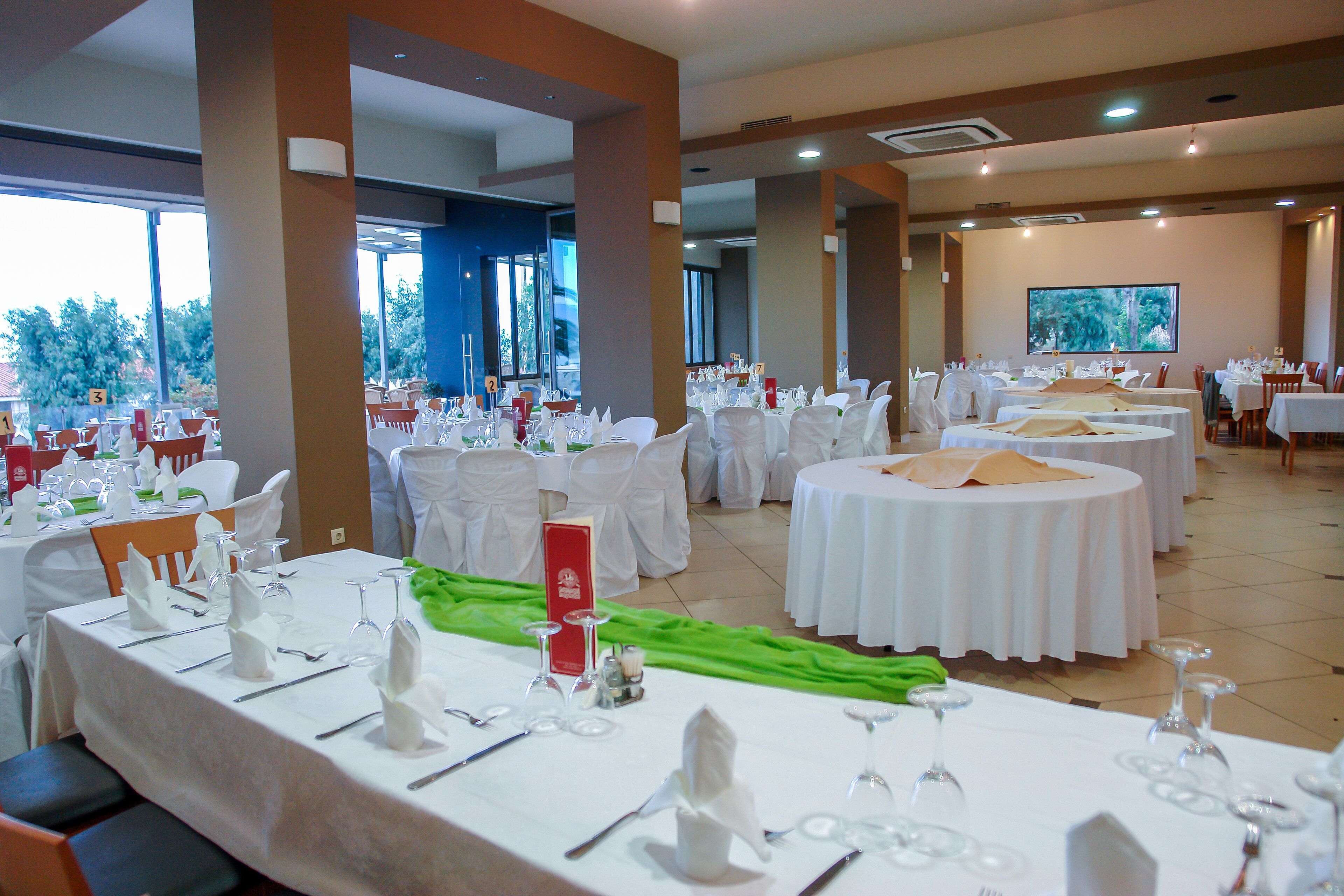 A banquet hall with round tables set for a formal event, large windows, and a view of trees.
