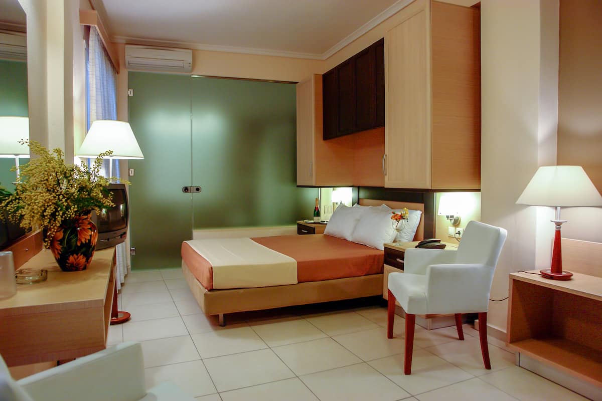 Una habitación de hotel con cama, mesita de noche, silla, lámpara y un amplio ventanal con cortinas.