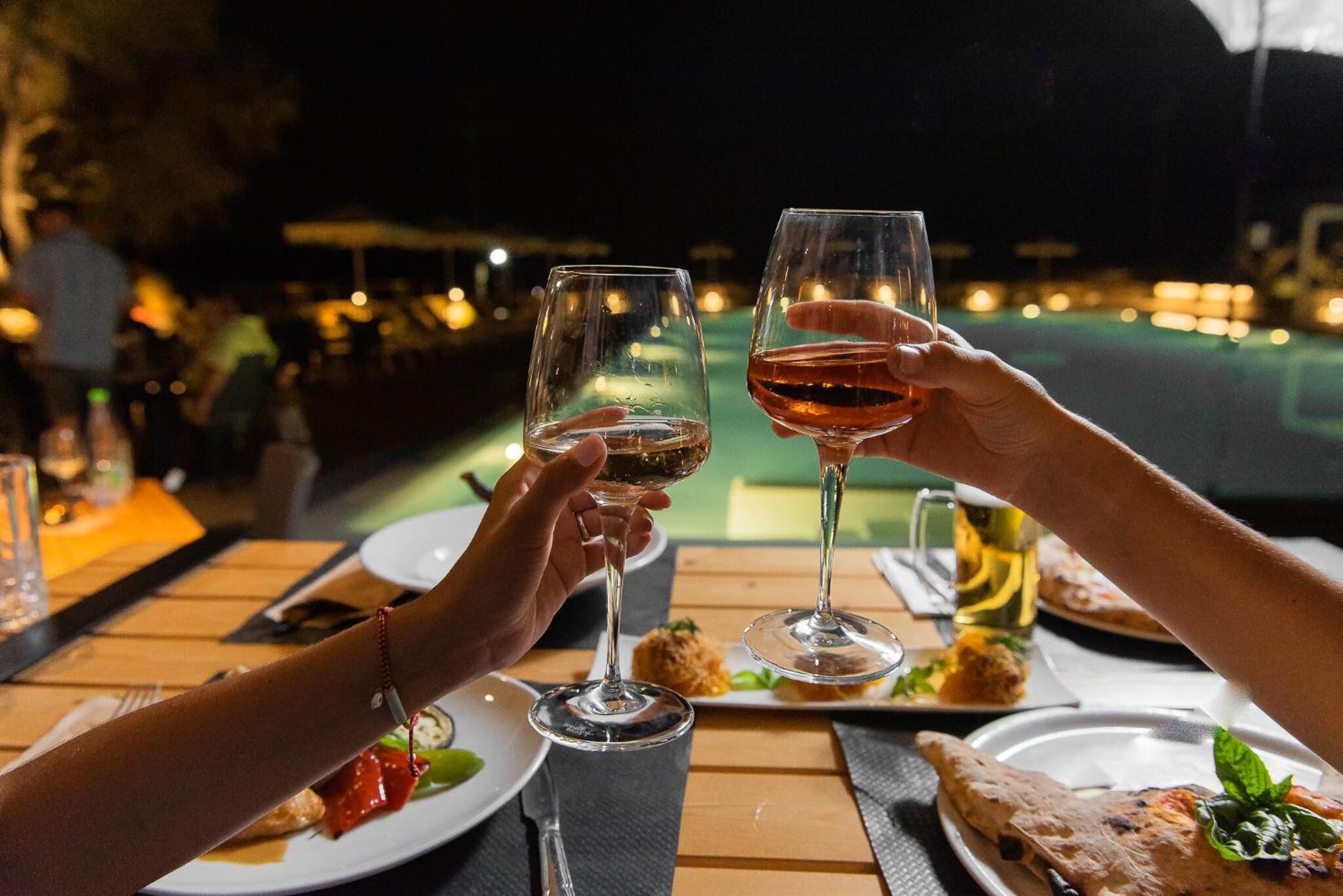 Two people toasting with wine glasses at a table with a pool view.