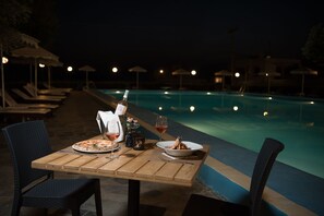 A dining table with a pizza, wine, and a candle at a poolside setting. - Marmari Bay Hotel (Karystos)