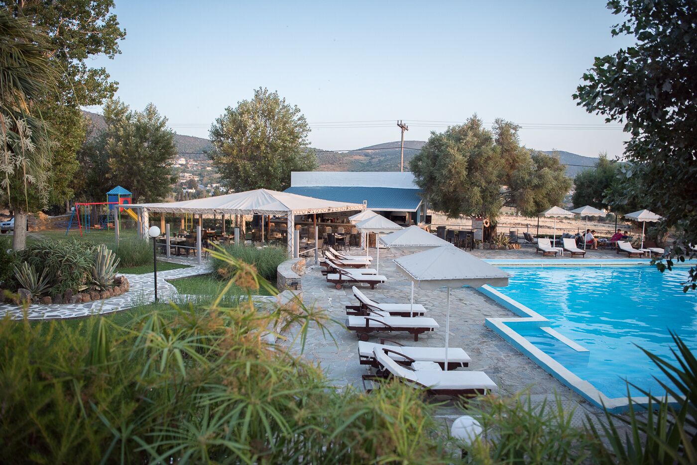 A poolside area with lounge chairs and umbrellas, a children's play area, and a building with a blue roof.