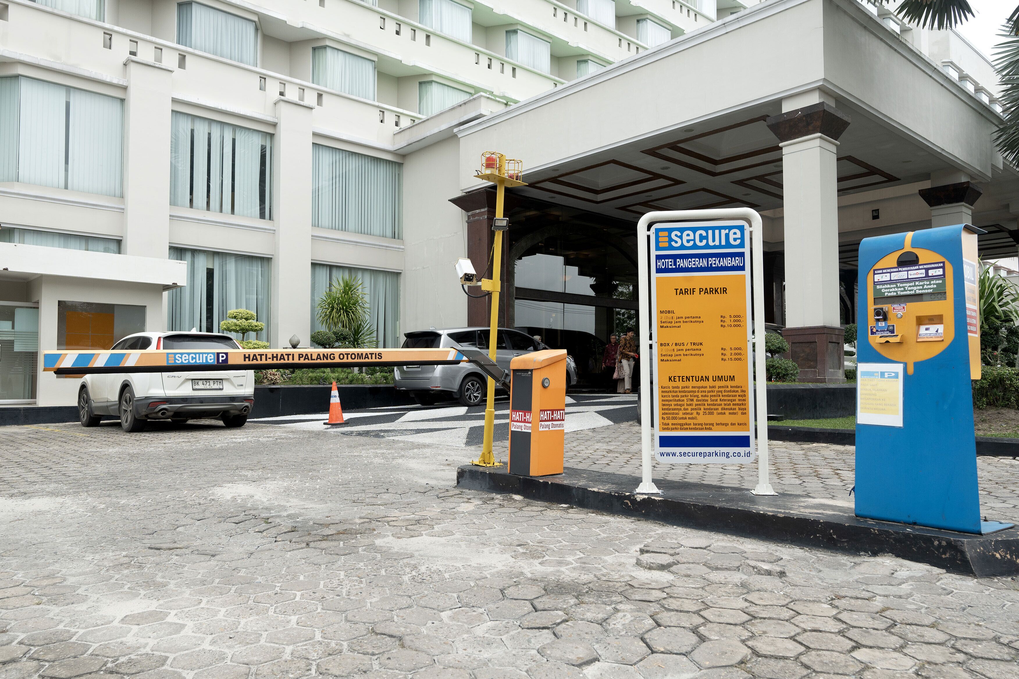 Entrada de hotel con punto de control de seguridad, un automóvil y un cartel que indica las tarifas de estacionamiento.