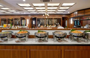 Een buffetopstelling met meerdere stations, waaronder schalen met rijst en andere gerechten, in een goed verlichte, moderne eetruimte met houten details en grote ramen.
