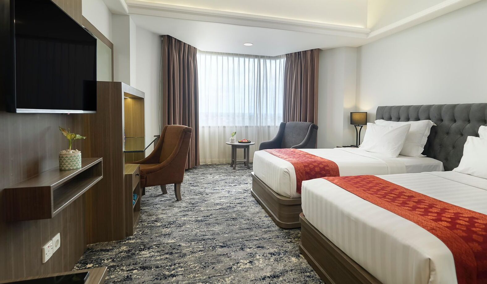 A hotel room with two beds, a desk, a chair, a TV, and a window with curtains.