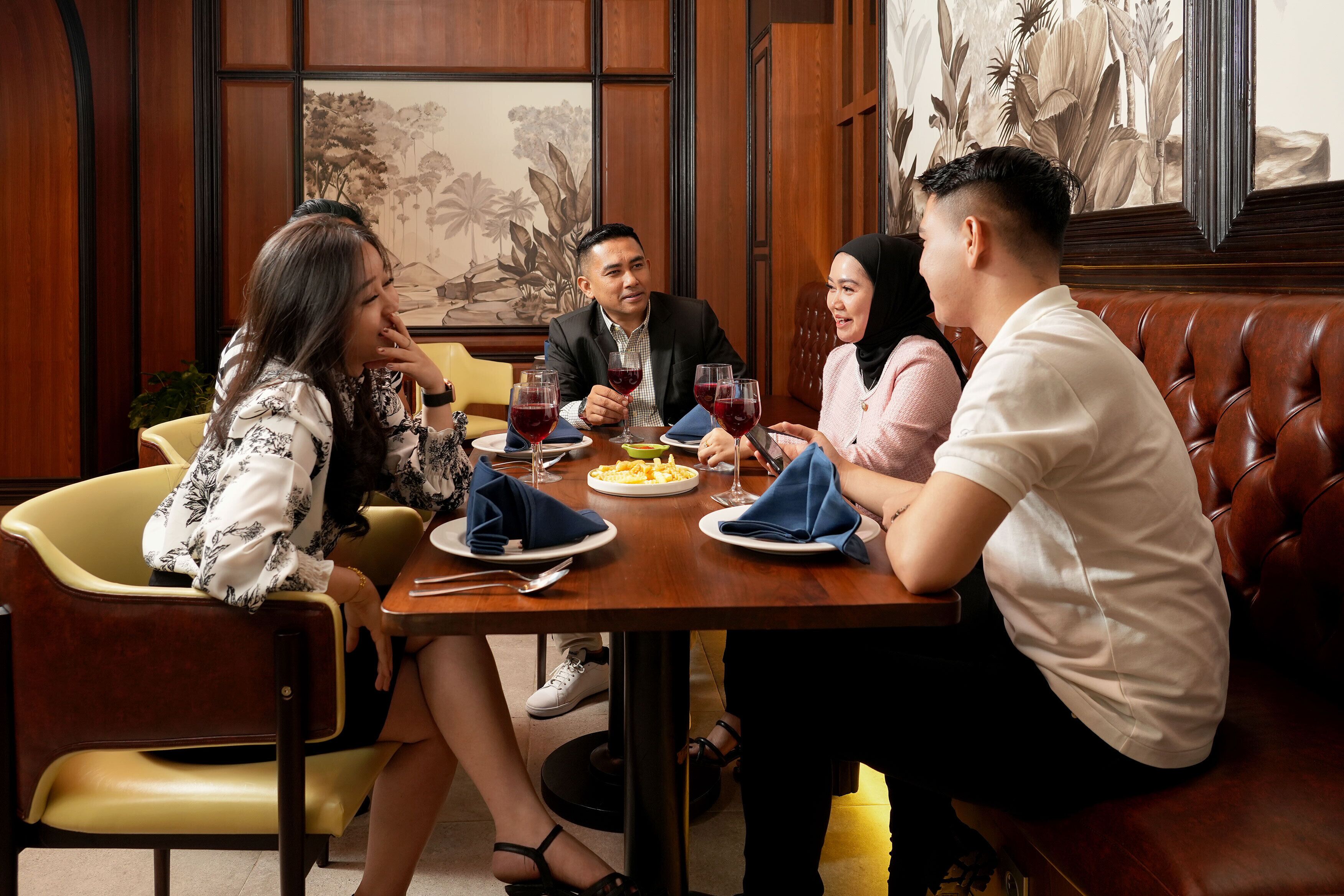 A group of people dining at a wooden table with a floral painting in the background.