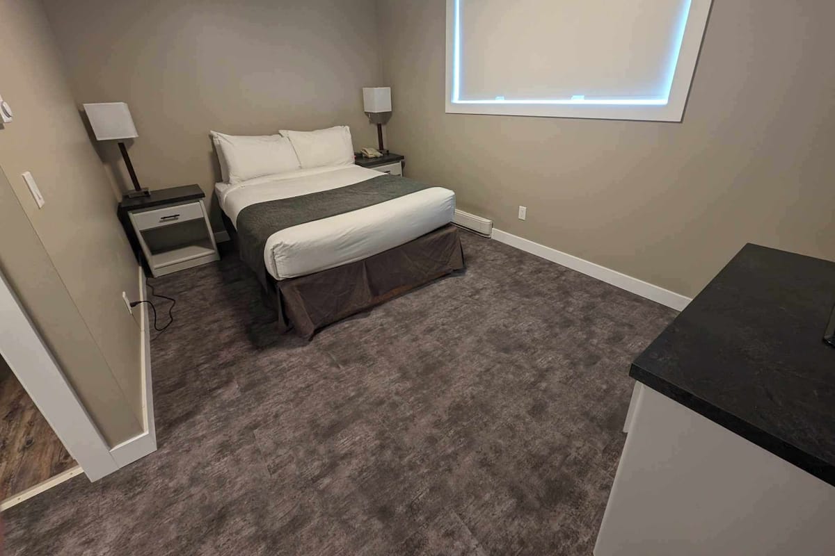 Une chambre à coucher comprenant un lit, des tables de chevet, un bureau, une fenêtre avec des stores et un sol recouvert d’une moquette.