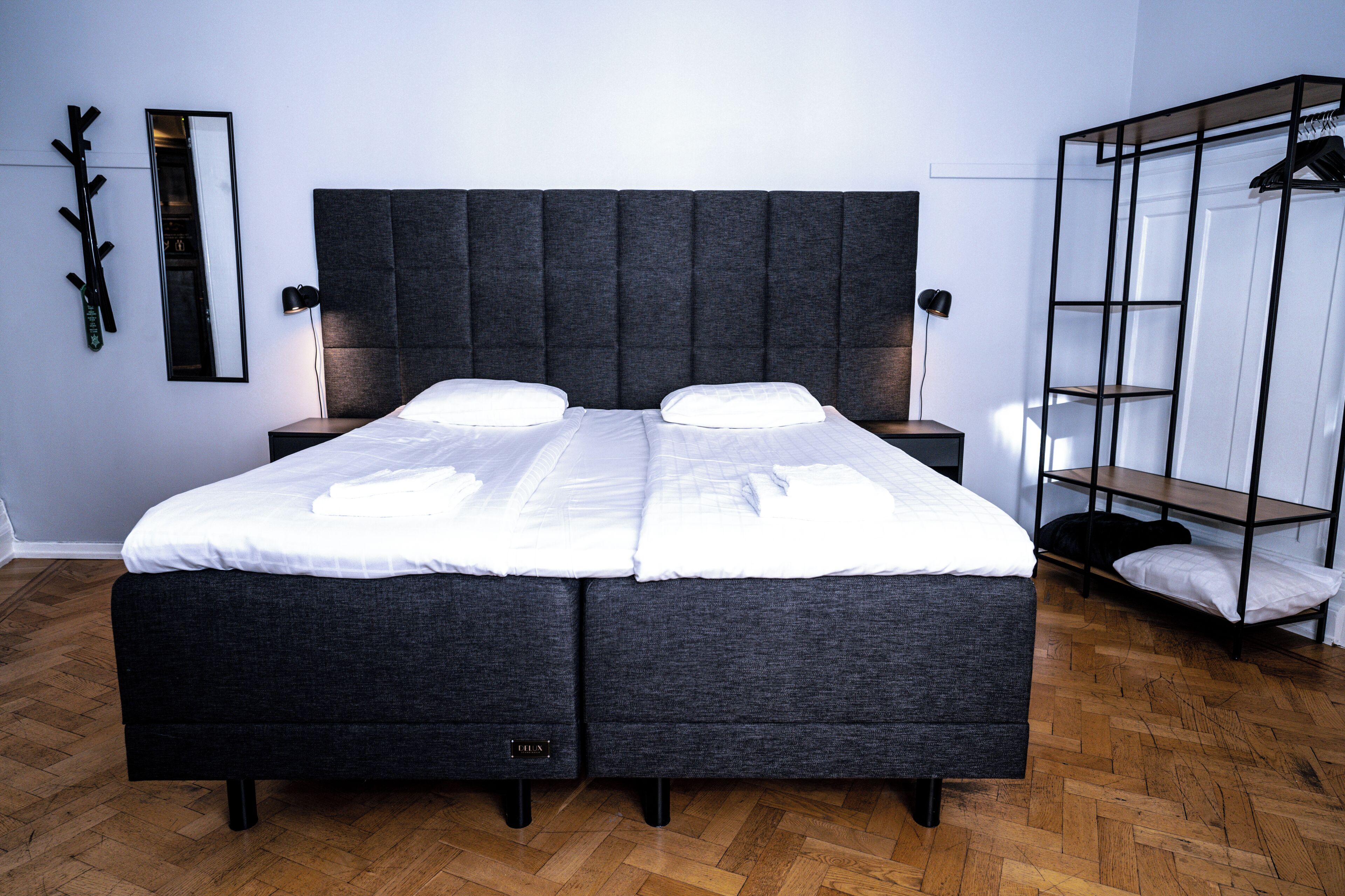 A double bed with a grey upholstered headboard, two bedside tables, a black hanger shelf, and a wooden herringbone floor.