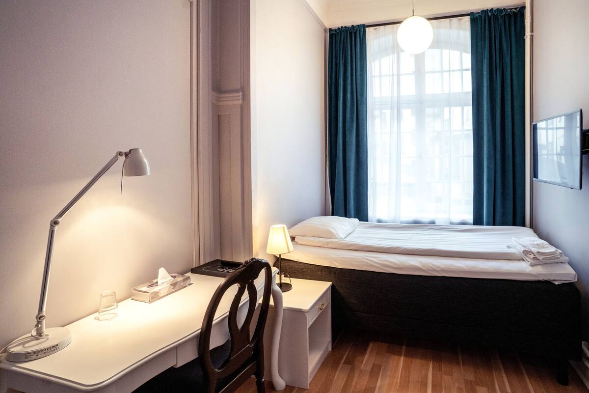 A hotel room with a bed, a desk with a lamp, a chair, and a window with curtains.