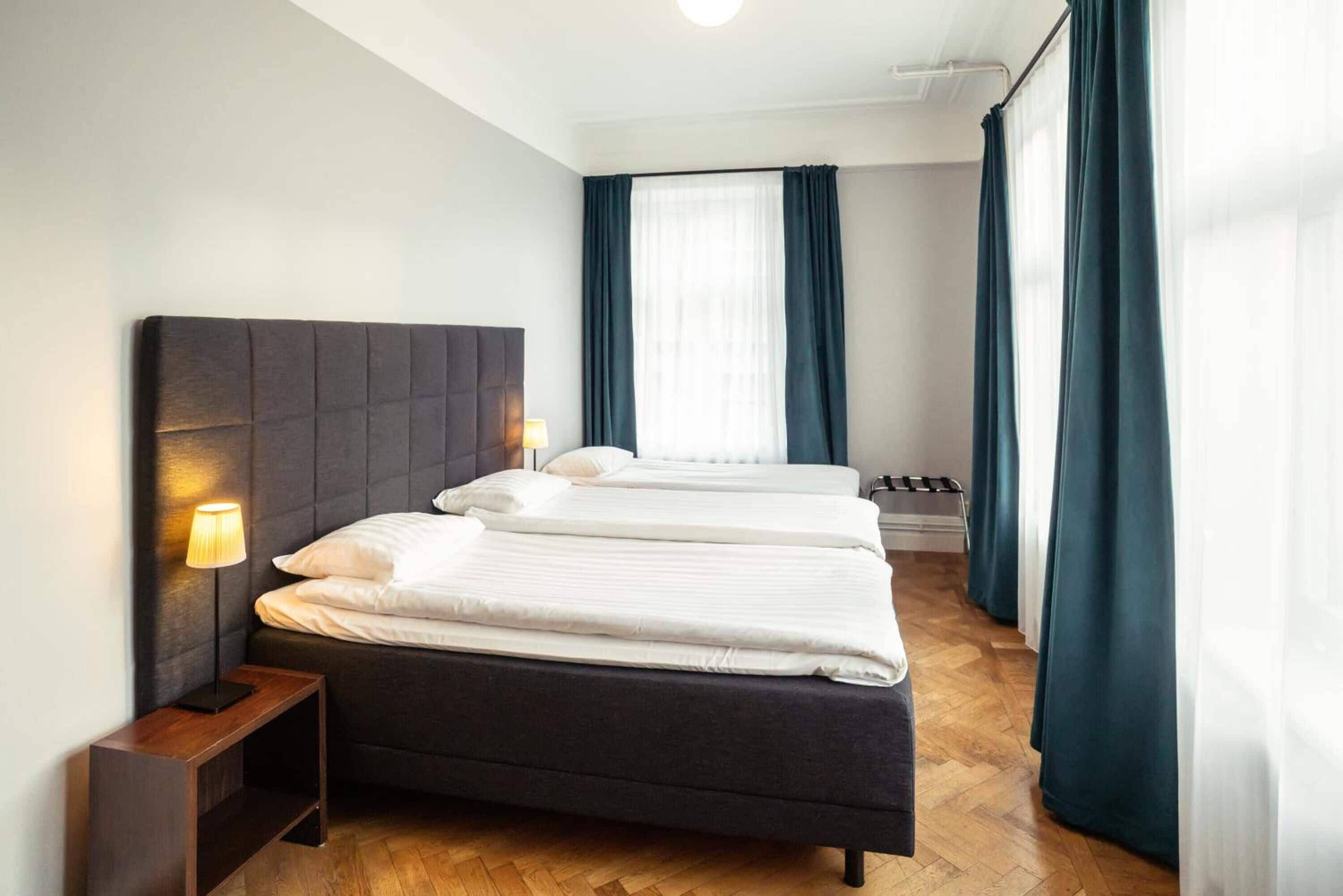 A hotel room with a dark headboard, two beds, a bedside table with a lamp, and a window with curtains.