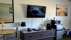A hotel room with a flat-screen TV mounted on the wall, a desk with a lamp, a microwave, and a refrigerator. There is a small plant on the desk and a painting on the wall. - Lord Nelson Motel (Kingston)