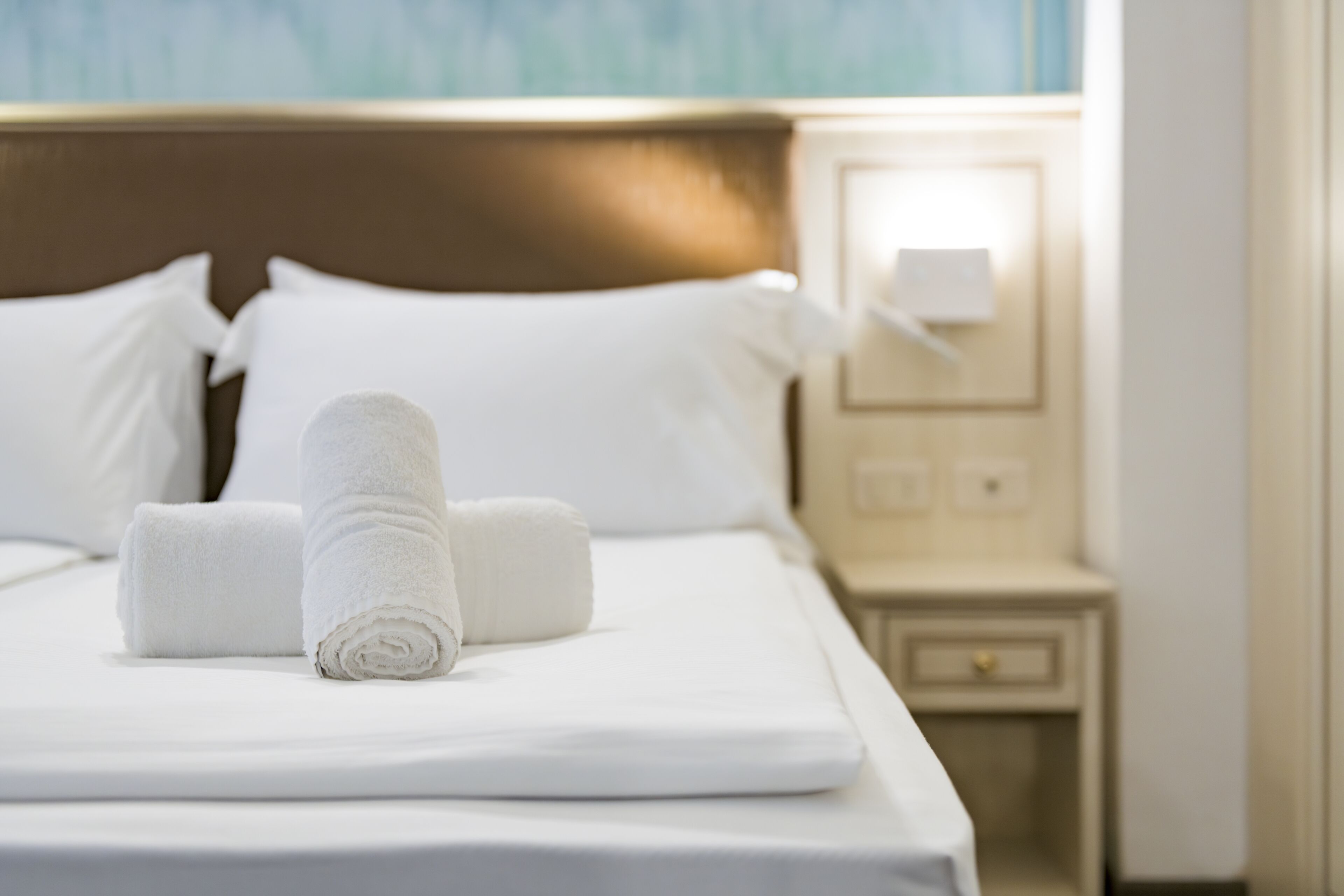 A neatly made bed with white linens and rolled towels, a wooden headboard, and a bedside table with a lamp.