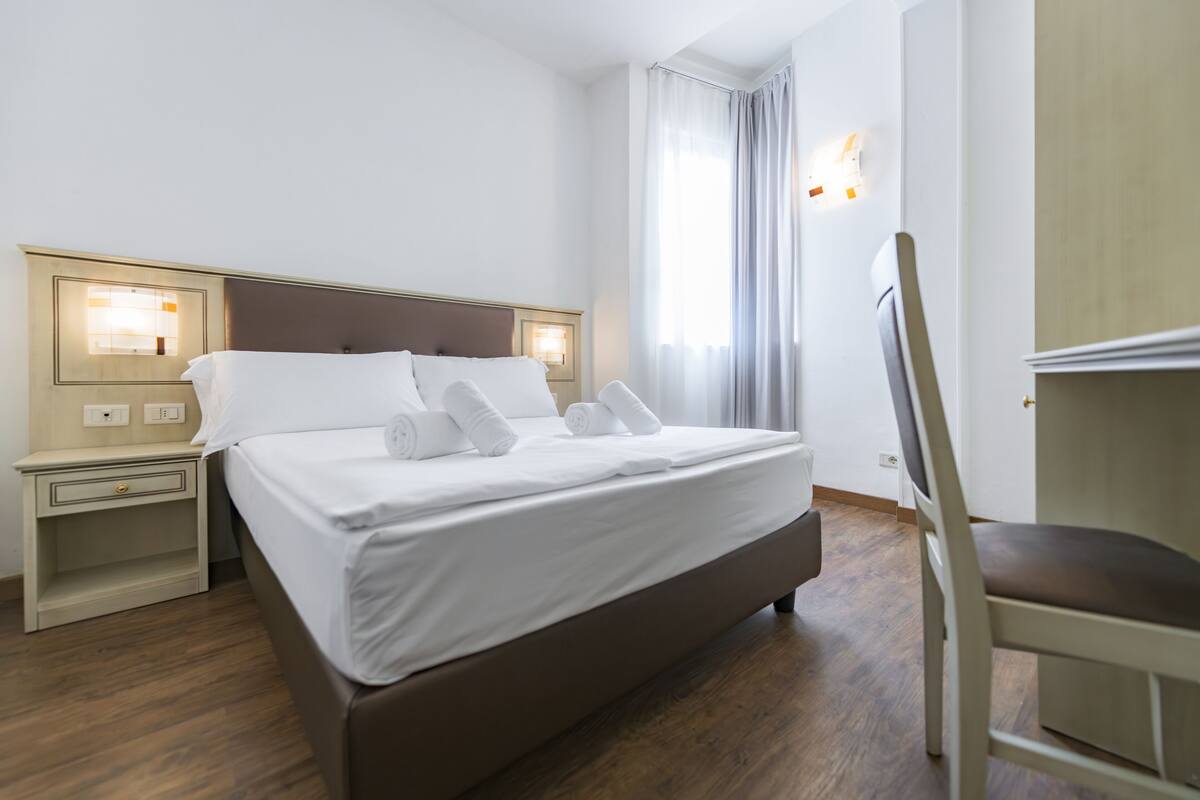 Een net opgemaakt bed met witte lakens en kussens, een nachtkastje met een lamp en een stoel.