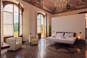 Amplia habitación de hotel con una cama grande, dos sillones, una mesita y un candelabro. La habitación cuenta con un techo alto con decoraciones ornamentales y ventanas amplias que ofrecen vistas al exterior.