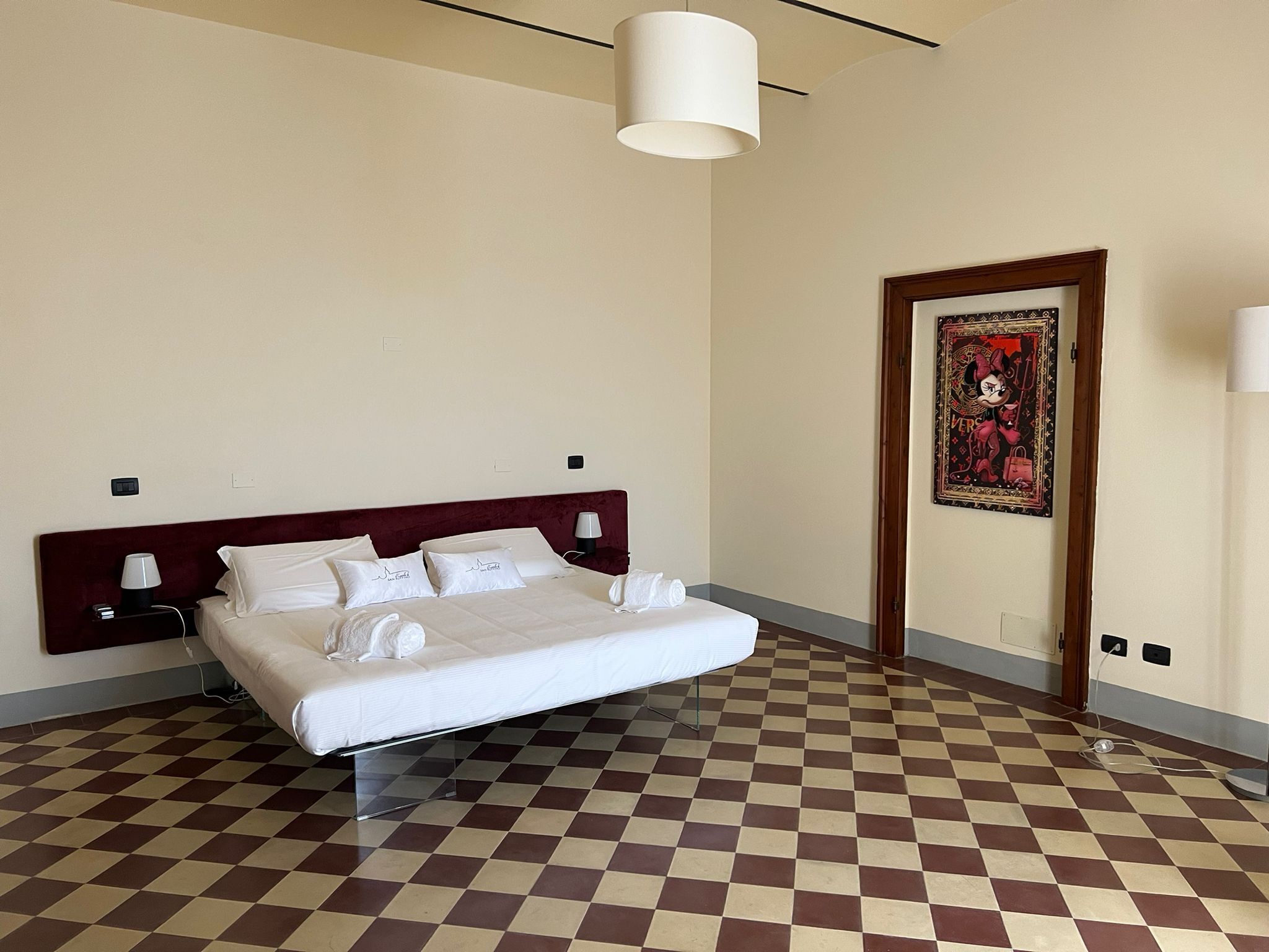 Una habitación de hotel con piso a cuadros, una cama grande con ropa blanca, un cabecero rojo y un cuadro en la pared.