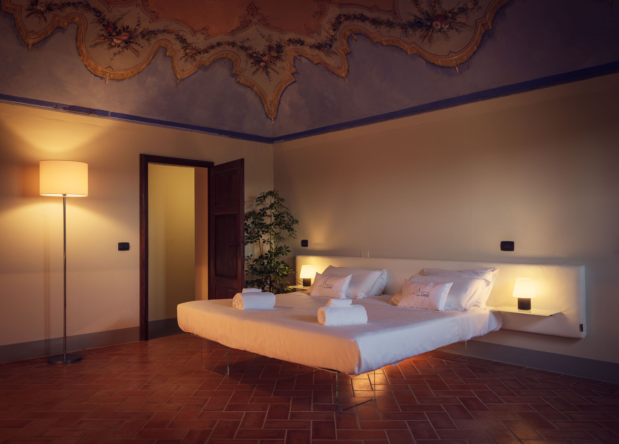 Habitación de hotel con una cama grande, una lámpara de pie, una planta en maceta y un techo decorativo.