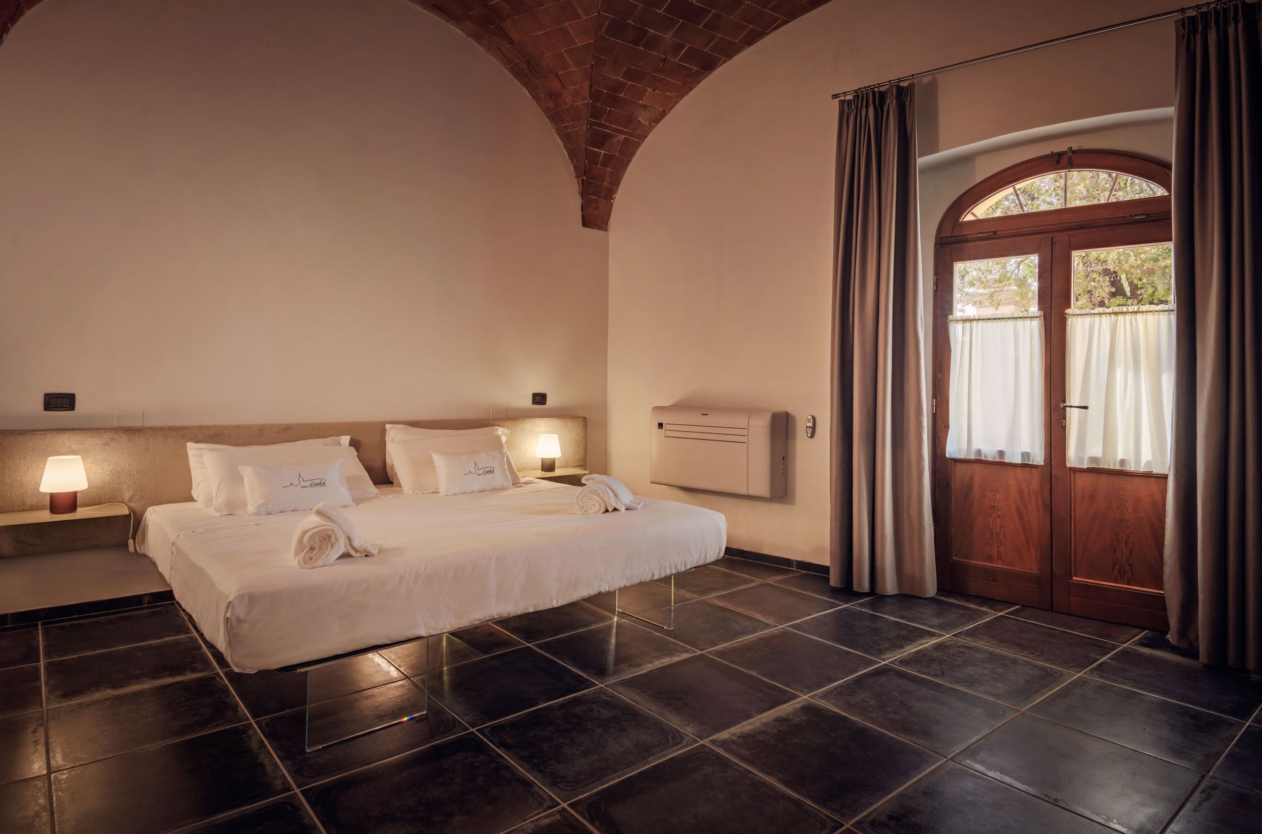Ein Hotelzimmer mit einem großen Bett, zwei Nachttischlampen, einer mit Vorhängen versehenen Holztür und einem gefliesten Boden.