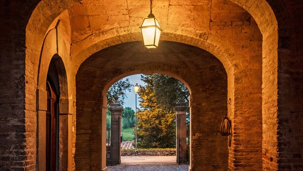 Een gewelfde stenen gang met een hanglamp, die uitkomt op een open gebied met bomen en een poort.