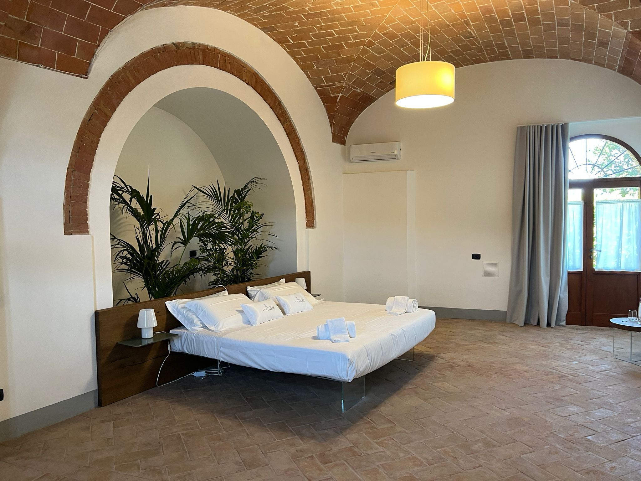 Ein Hotelzimmer mit Bett, einer Topfpflanze, einer Pendelleuchte und einem Fenster mit Vorhängen.