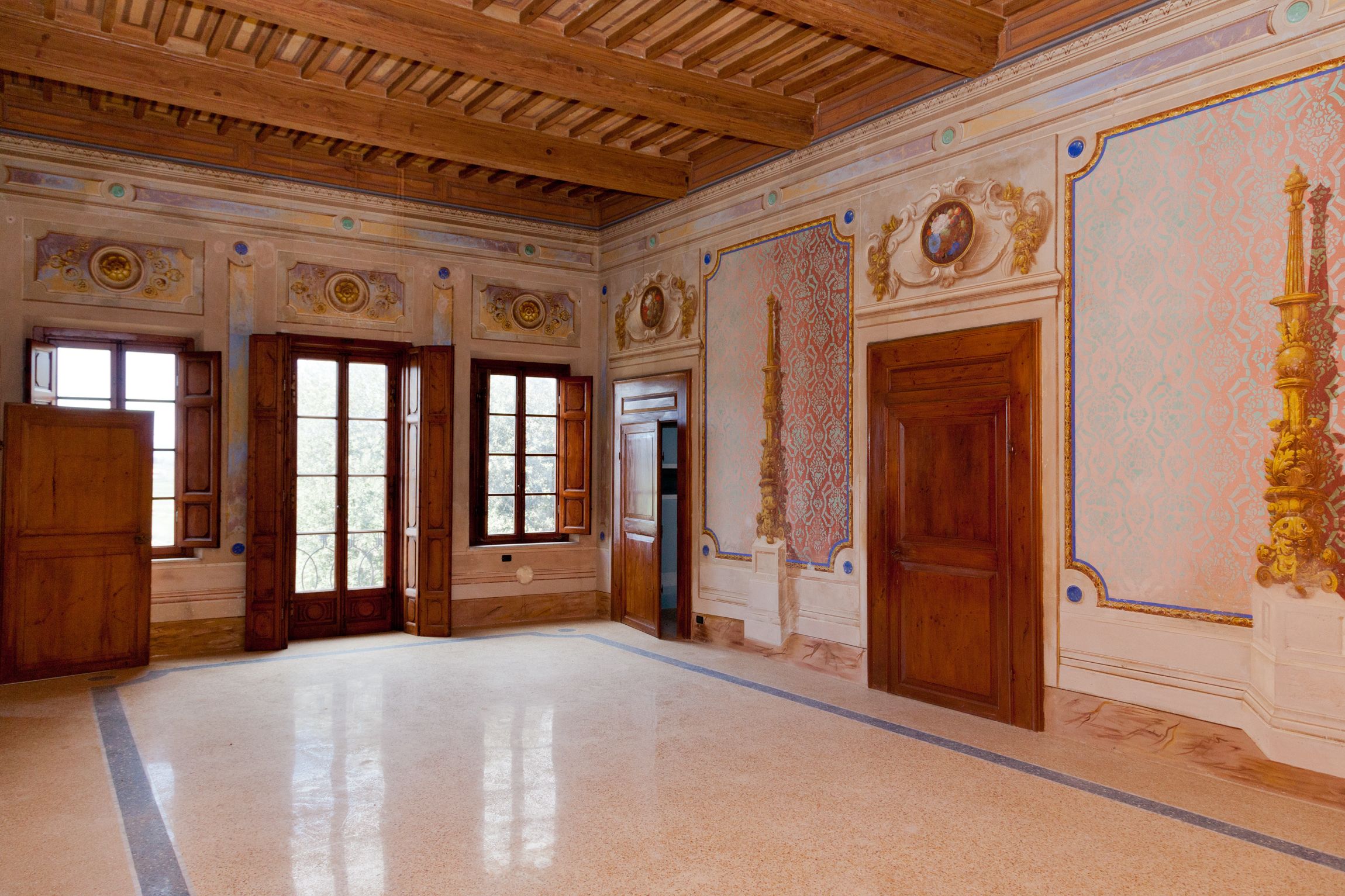 Una habitación amplia con puertas de madera, ventanas grandes y decoraciones ornamentales en las paredes.