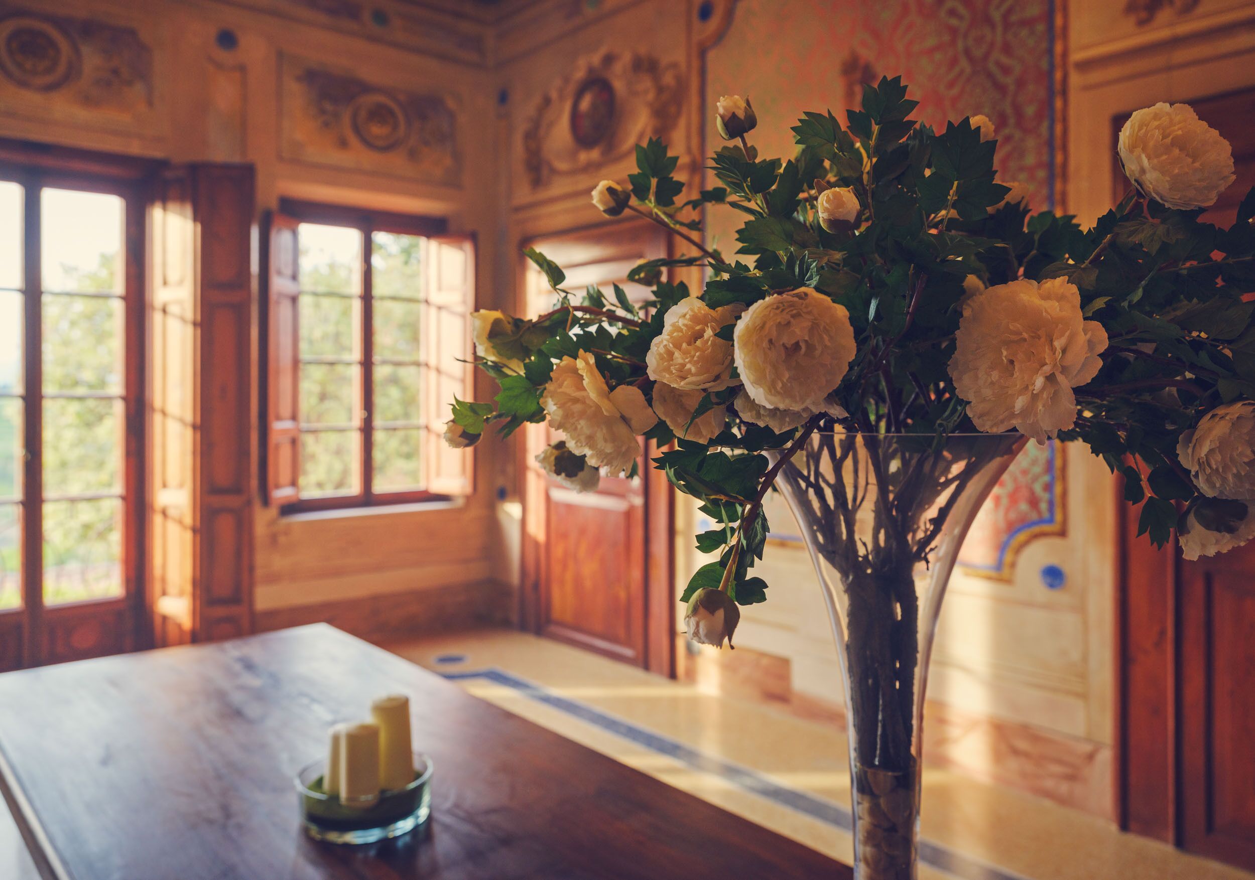 A room with a wooden table, a vase with white flowers, and a window with a view of greenery.