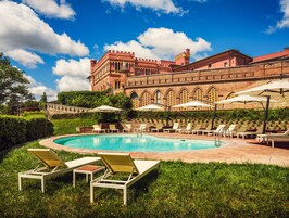 Una piscina con sillones de descanso y sombrillas, rodeada de un césped cuidado y un edificio histórico grande con ventanas en arco y una torre.