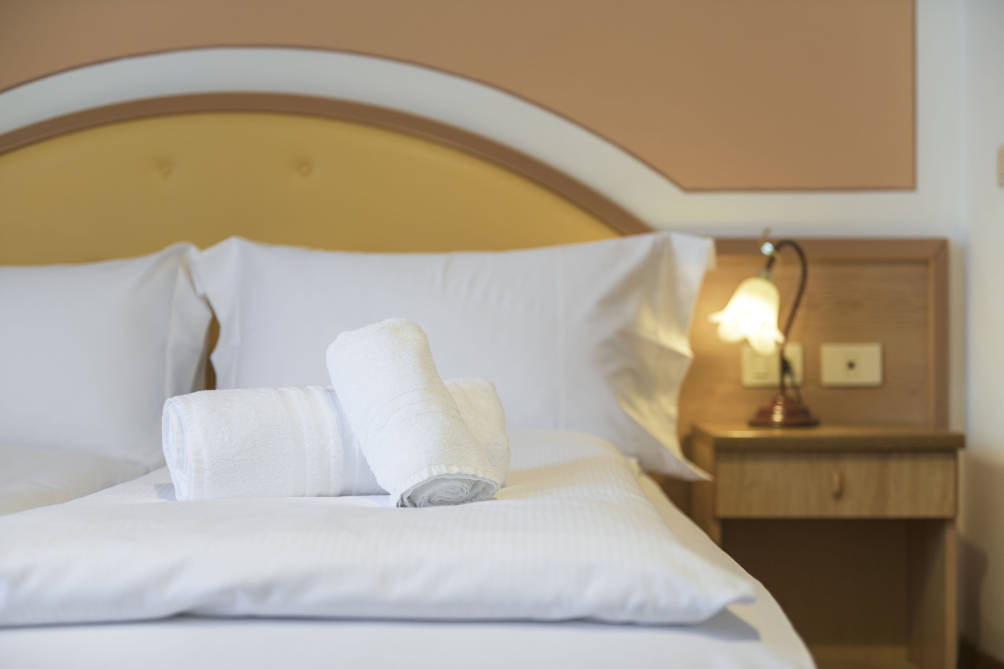Un lit bien fait, avec du linge de lit blanc et des serviettes pliées, une tête de lit en bois et une table de chevet avec une lampe.