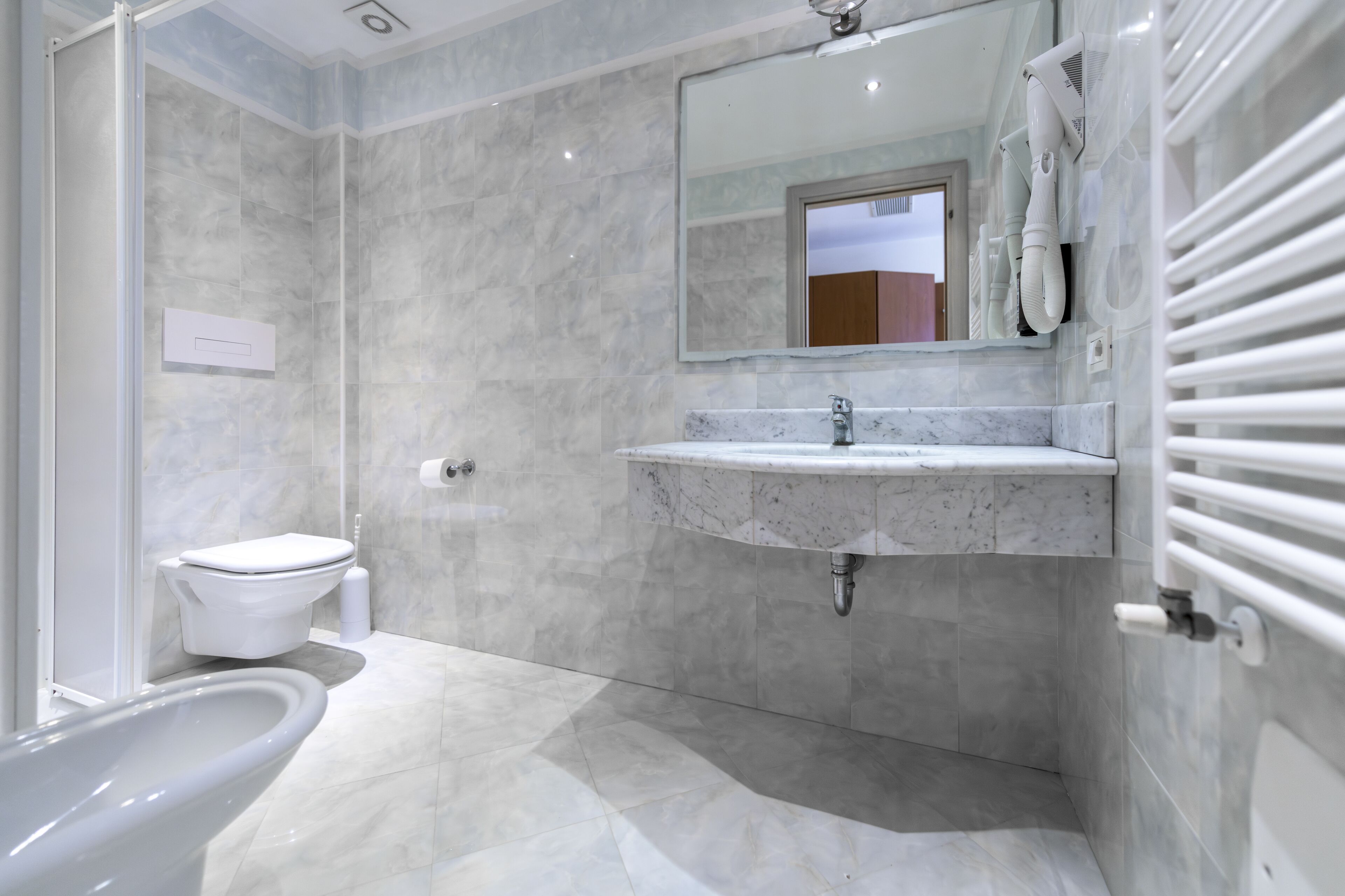 Un baño moderno con piso de mármol, inodoro blanco, lavamanos con espejo y zona de ducha.