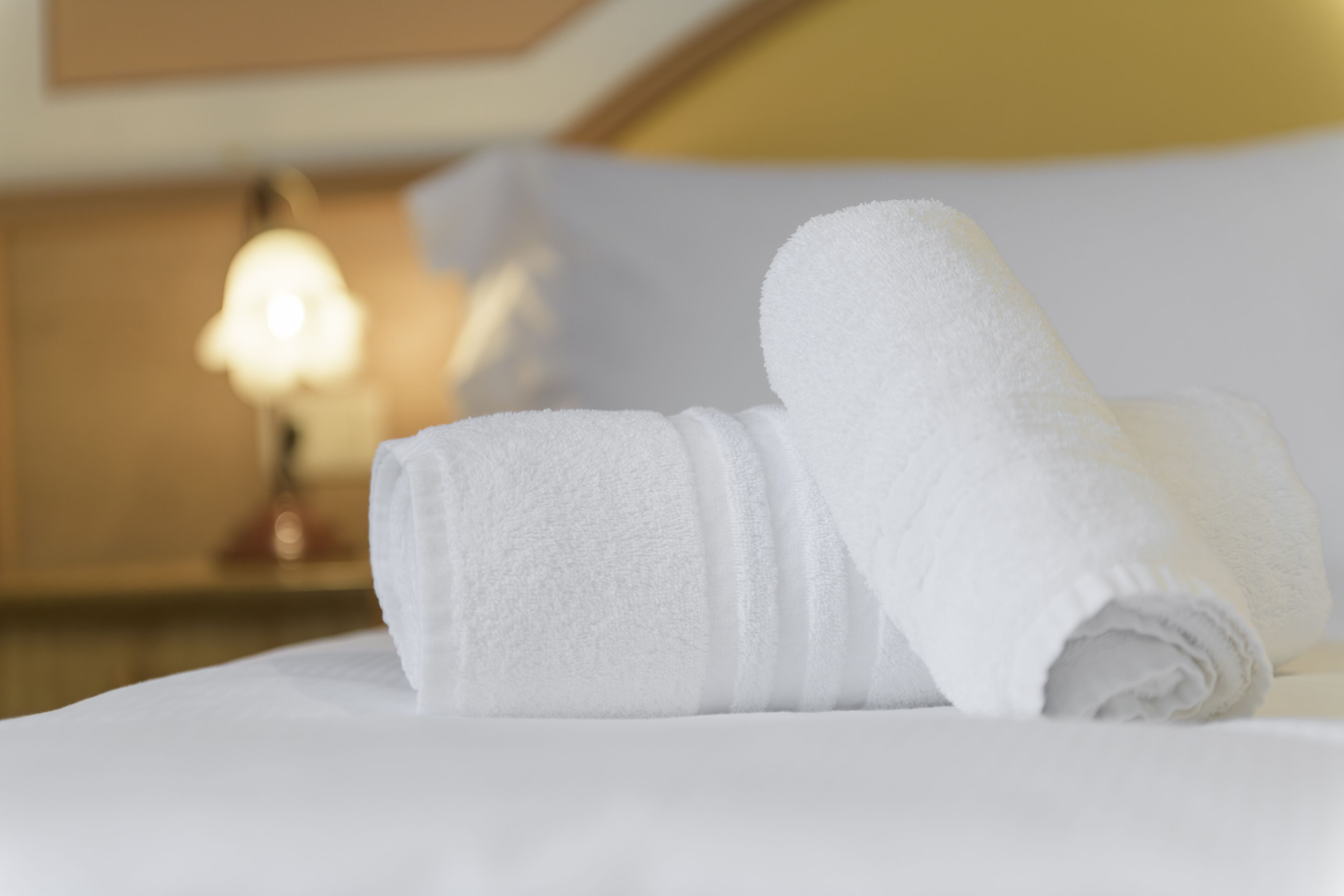 A neatly made bed with white linens and a rolled white towel on top.