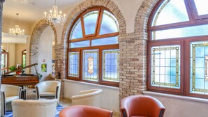 A spacious lobby with arched brick walls, large windows with stained glass, a grand piano, leather armchairs, and a chandelier.