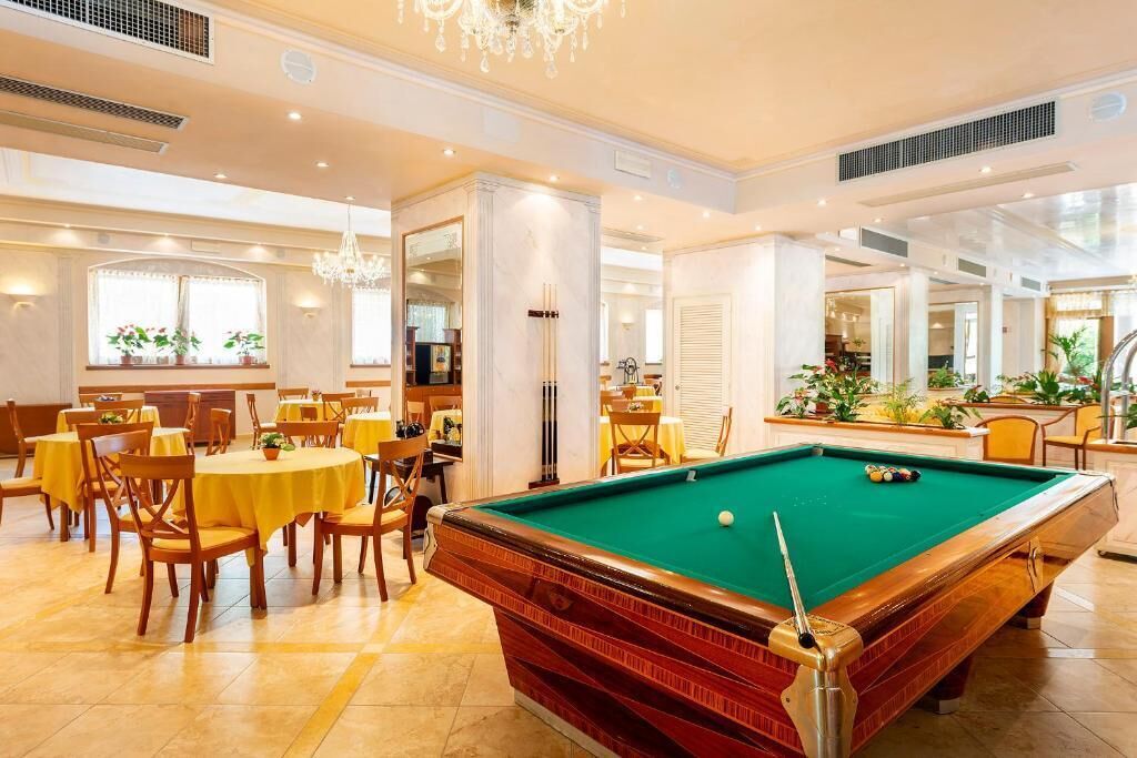A billiards room with a pool table, dining area with yellow tables and chairs, and a chandelier.
